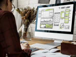 Person working at a desk on a computer displaying website wireframes and layout sketches, illustrating the strategic planning phase of a website redesign process.