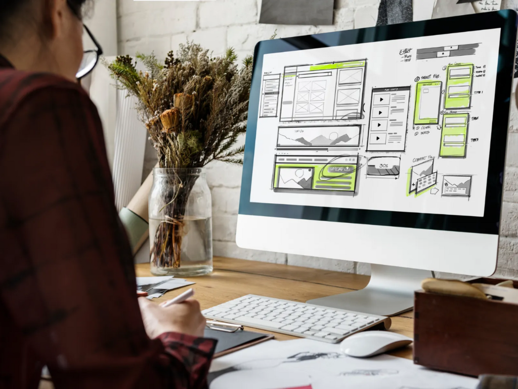 Person working at a desk on a computer displaying website wireframes and layout sketches, illustrating the strategic planning phase of a website redesign process.