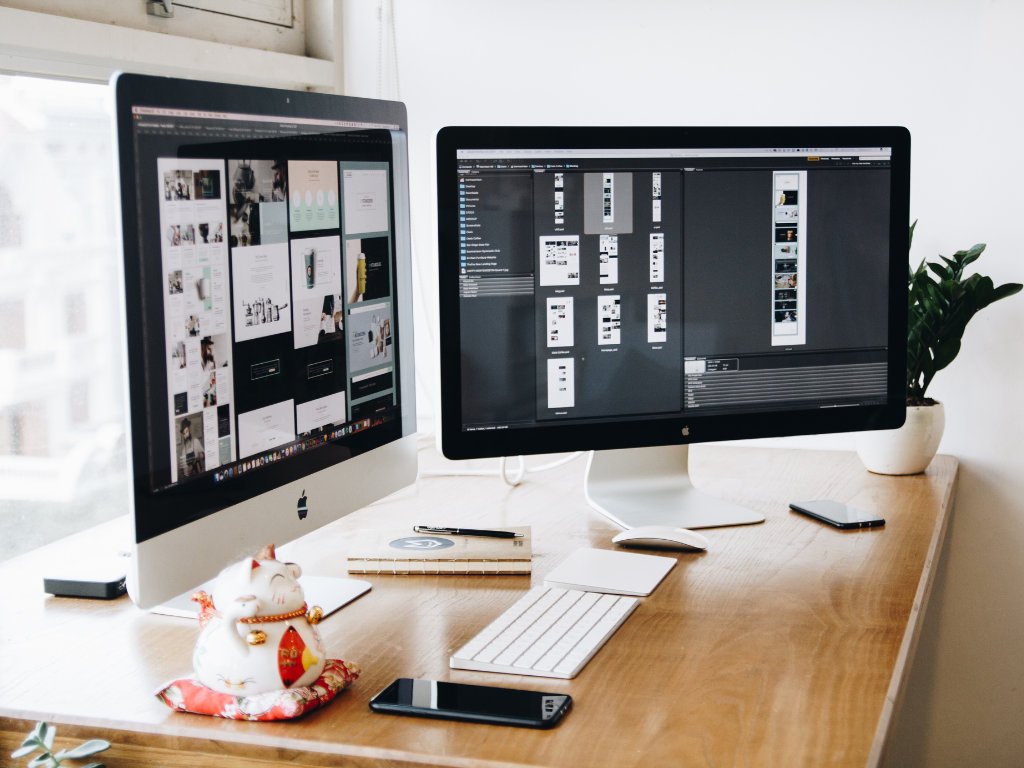Workspace with two large computer monitors displaying web design layouts on a wooden desk with a keyboard, phone, notebook, and small plant.
